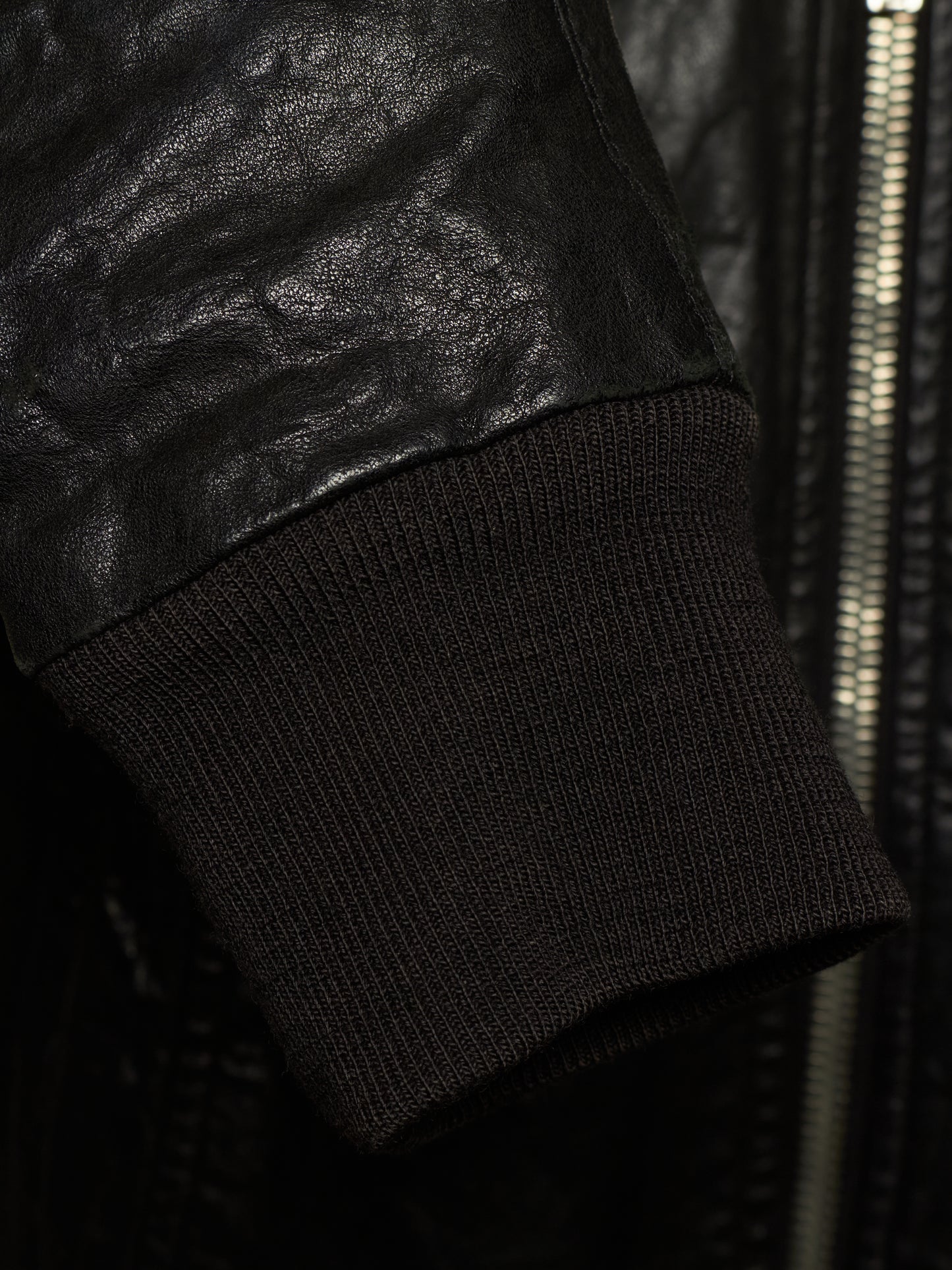Close-up detail of a black leather jacket sleeve meeting a black ribbed knit cuff, with a zipper visible in the background.