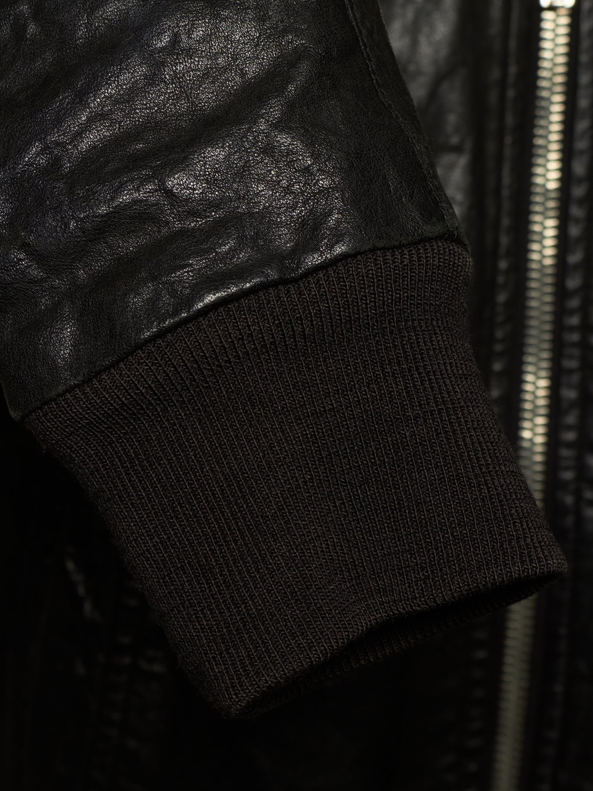 Close-up detail of a black leather jacket sleeve meeting a black ribbed knit cuff, with a zipper visible in the background.