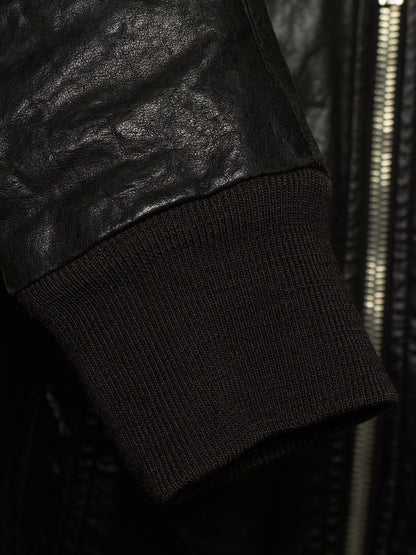 Close-up detail of a black leather jacket sleeve meeting a black ribbed knit cuff, with a zipper visible in the background.