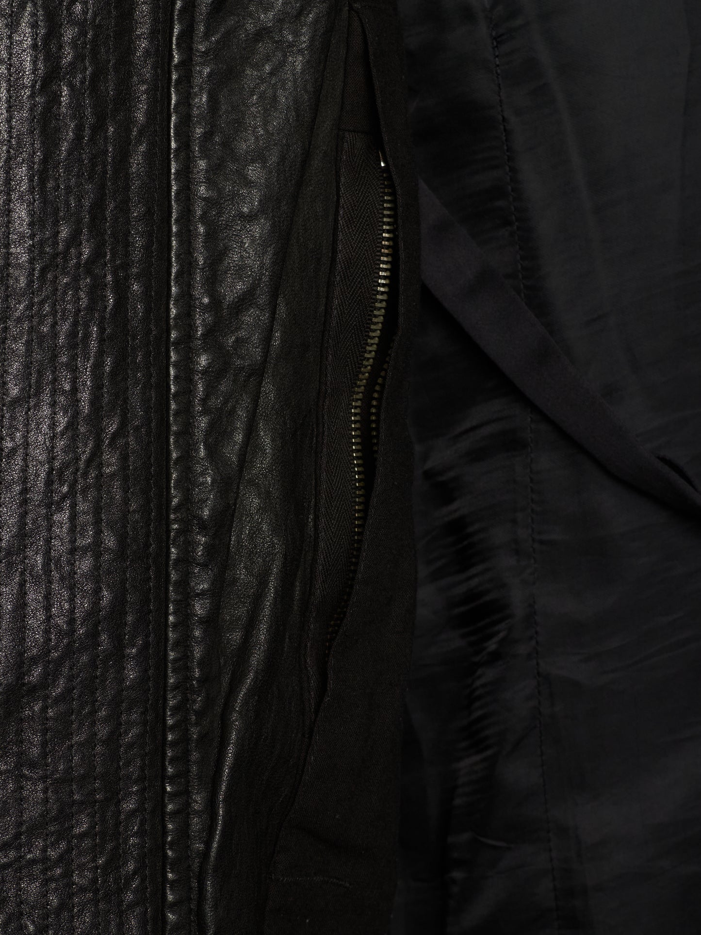Close-up detail of a black jacket showing a textured leather-like panel, a black zipper, and a smooth black fabric interior.