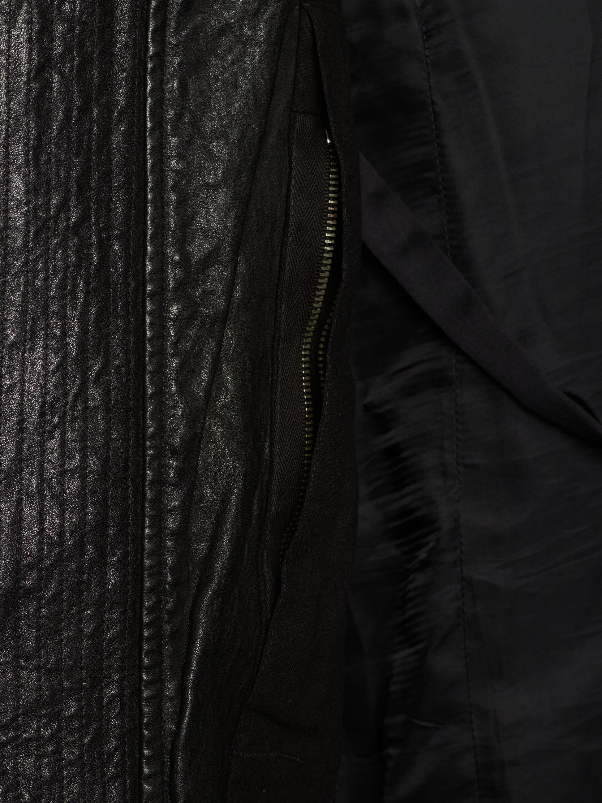 Close-up detail of a black jacket showing a textured leather-like panel, a black zipper, and a smooth black fabric interior.
