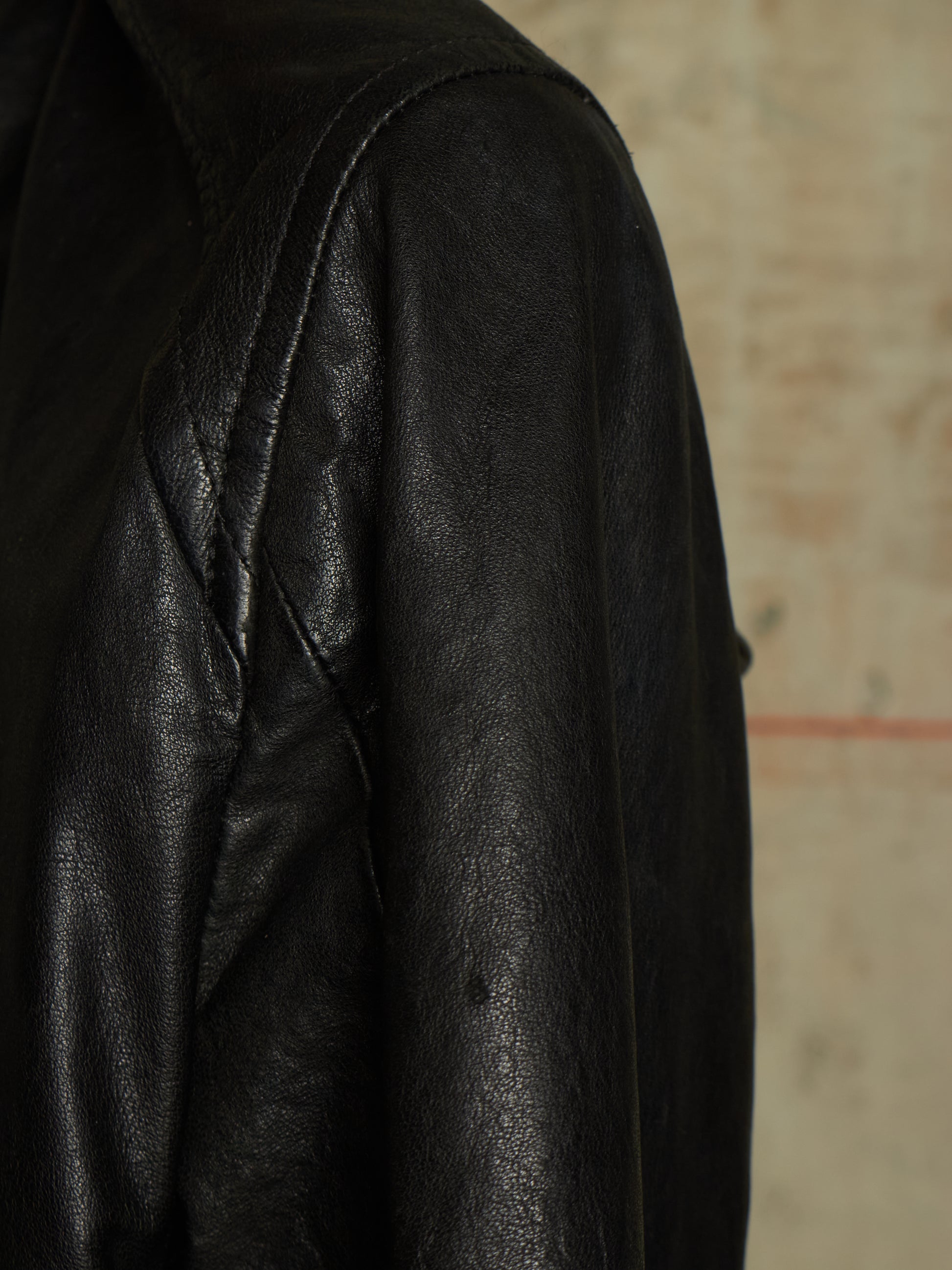 Close-up of a black leather jacket showing the textured leather fabric and shoulder seam against a neutral beige background.