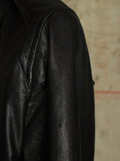 Close-up of a black leather jacket showing the textured leather fabric and shoulder seam against a neutral beige background.