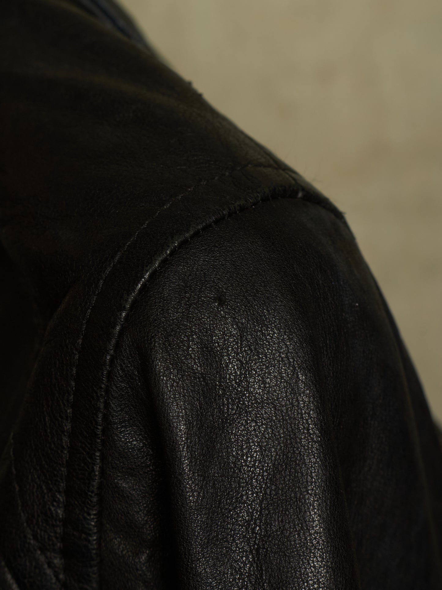 Close-up detail of a black leather jacket showing the textured leather fabric and shoulder seam against a neutral beige background.