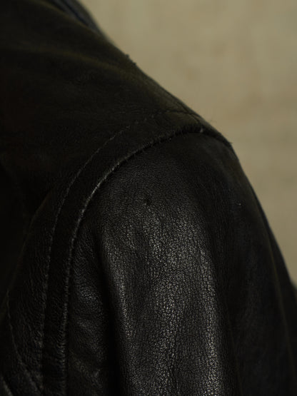 Close-up detail of a black leather jacket showing the textured leather fabric and shoulder seam against a neutral beige background.