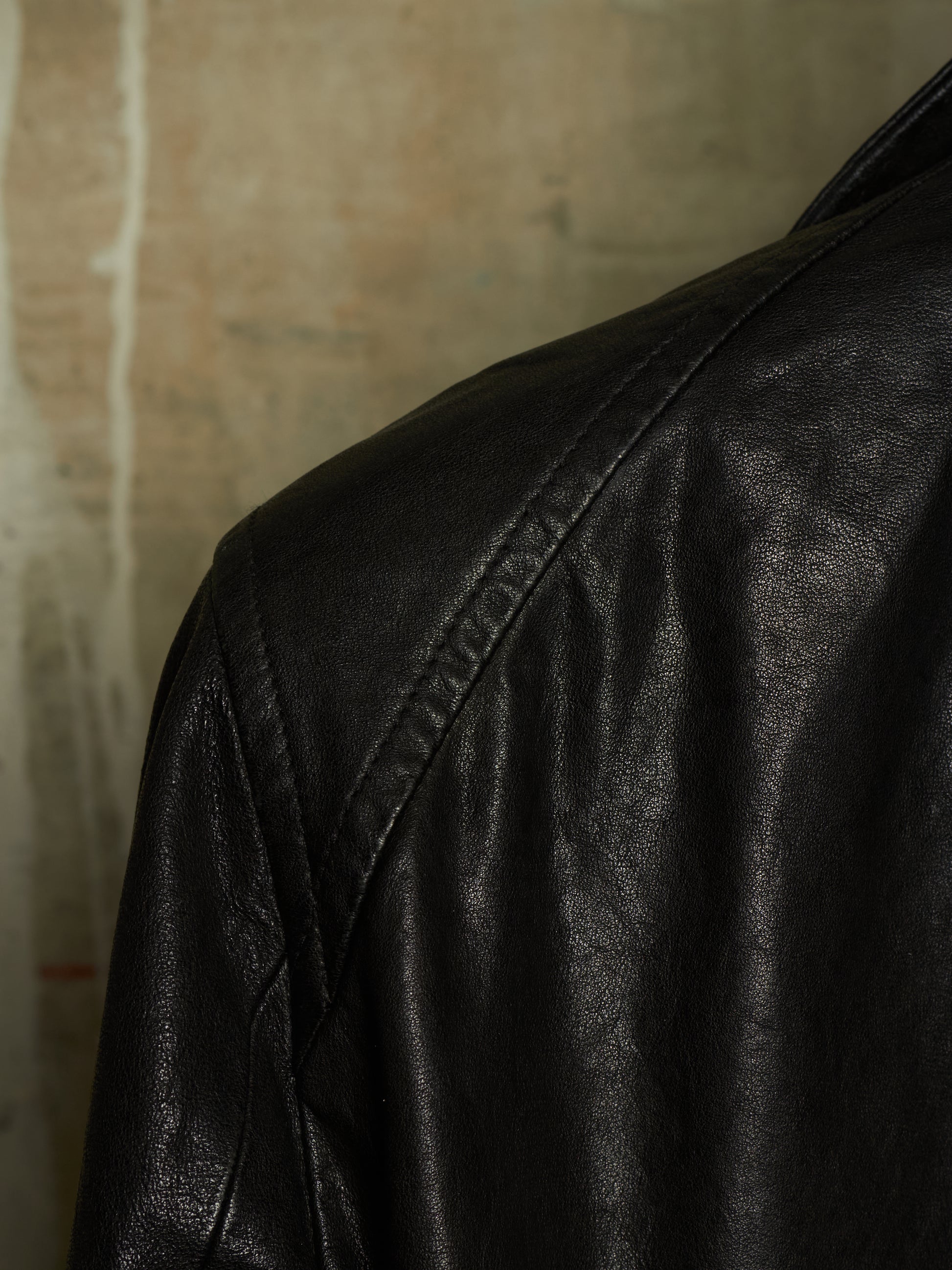 Close-up of a black leather jacket showing the shoulder and upper back with visible stitching against a textured beige background.