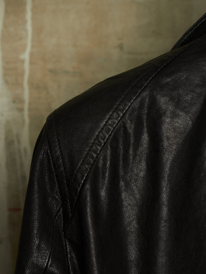 Close-up of a black leather jacket showing the shoulder and upper back with visible stitching against a textured beige background.