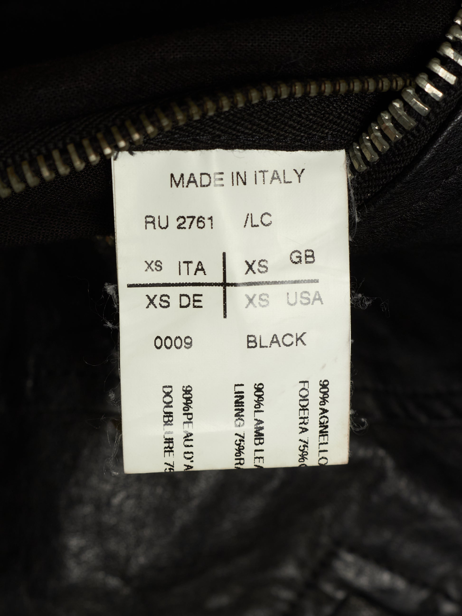 A close-up of a white size and material label sewn inside a black garment near the zipper. The label reads 'MADE IN ITALY', shows size information in Italian, English, German, and US, and indicates the fabric is 90% Angello and 70% polyamide, with 'BLACK' printed at the bottom.