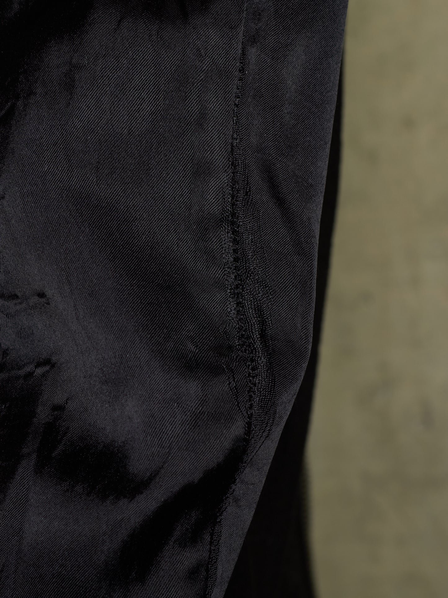 Close-up detail of a black textured fabric garment, showing a seam and weave, with a blurred beige background on the right.