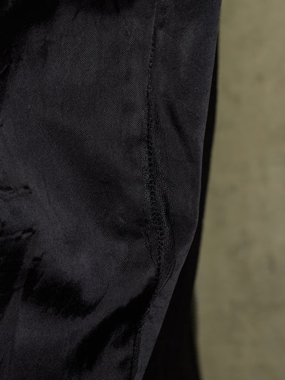 Close-up detail of a black textured fabric garment, showing a seam and weave, with a blurred beige background on the right.