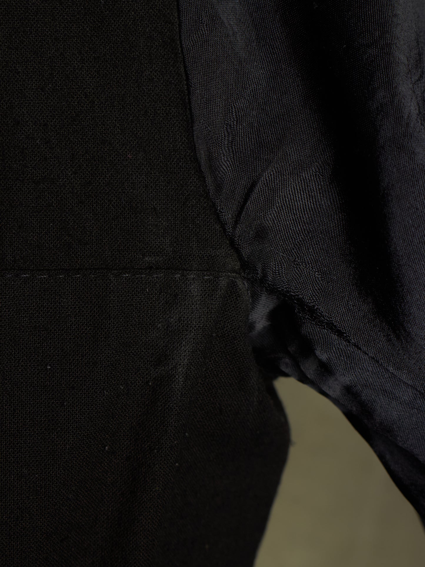 Close-up detail of a black textured fabric garment, showing a seam and the side of a sleeve or body section against a dark background.