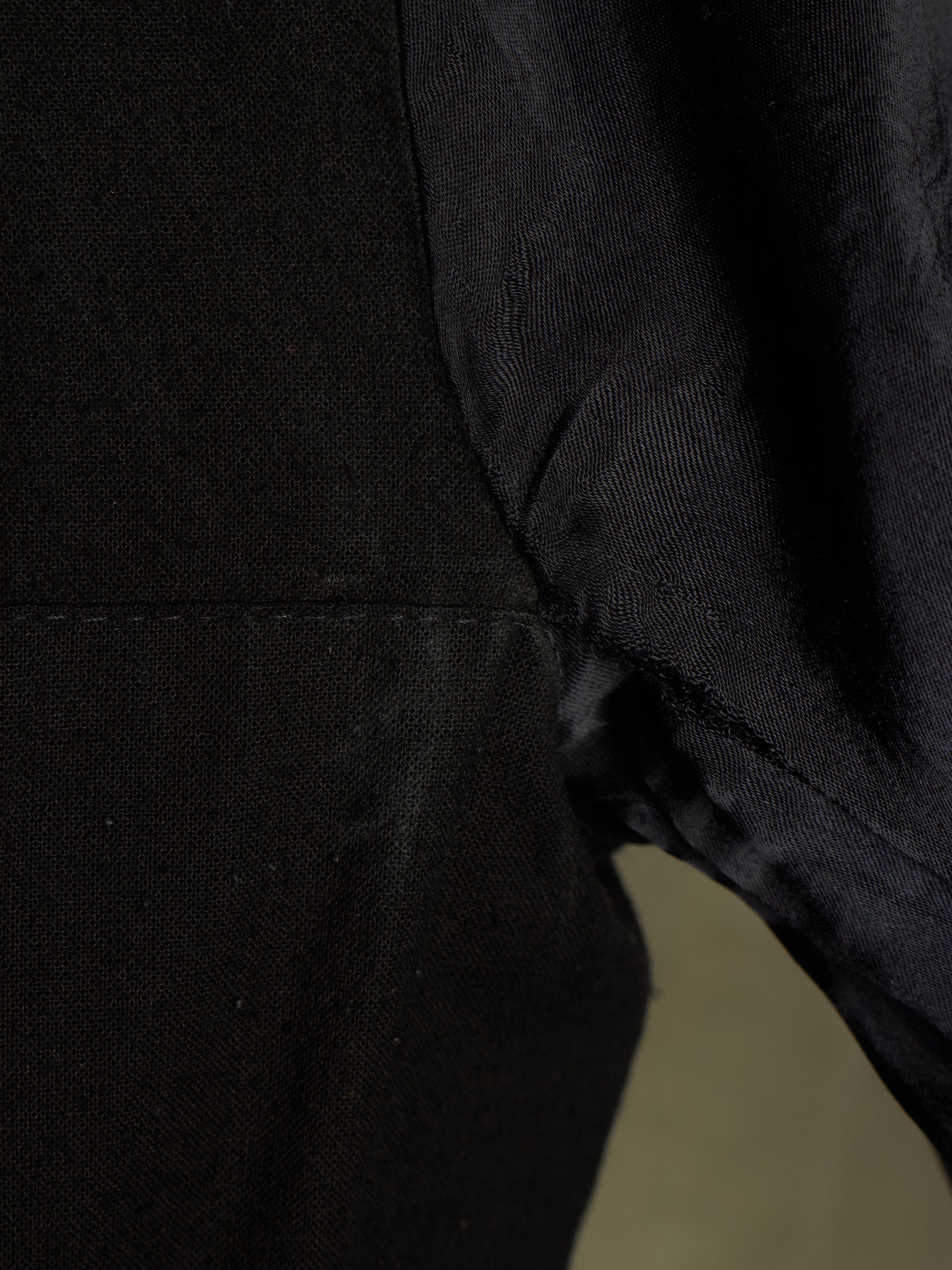 Close-up detail of a black textured fabric garment, showing a seam and the side of a sleeve or body section against a dark background.
