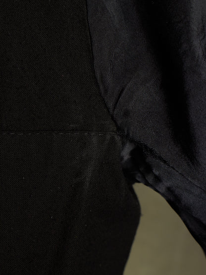 Close-up detail of a black textured fabric garment, showing a seam and the side of a sleeve or body section against a dark background.