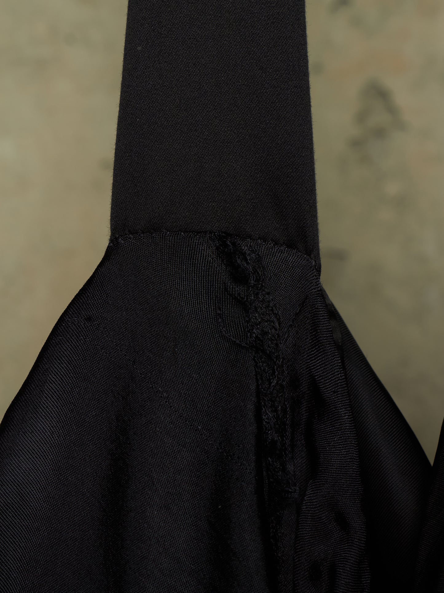 Close-up detail of a black fabric bag or garment showing a gathered seam, textured woven fabric, and a small tassel detail against a neutral beige background.