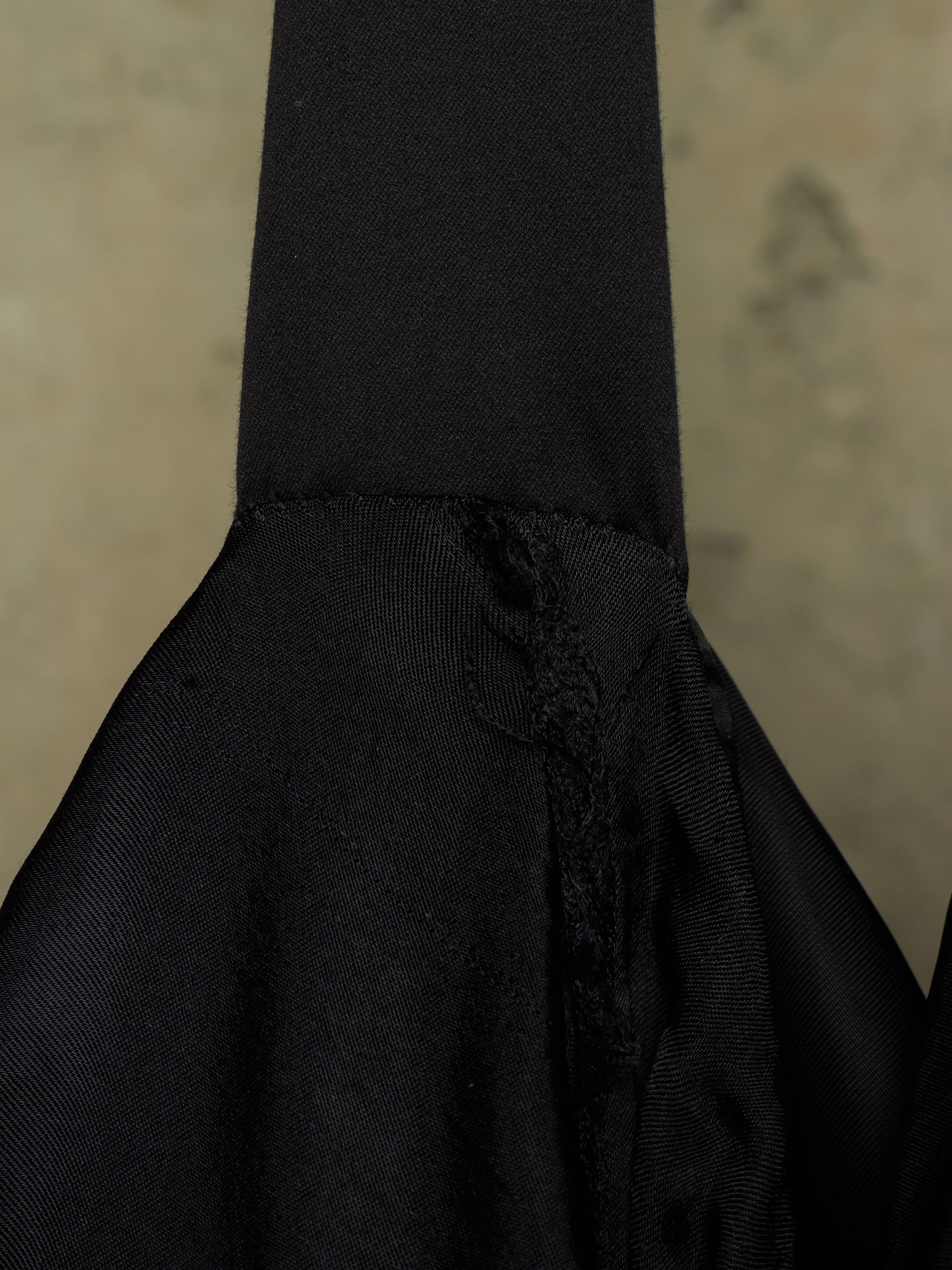 Close-up detail of a black fabric bag or garment showing a gathered seam, textured woven fabric, and a small tassel detail against a neutral beige background.