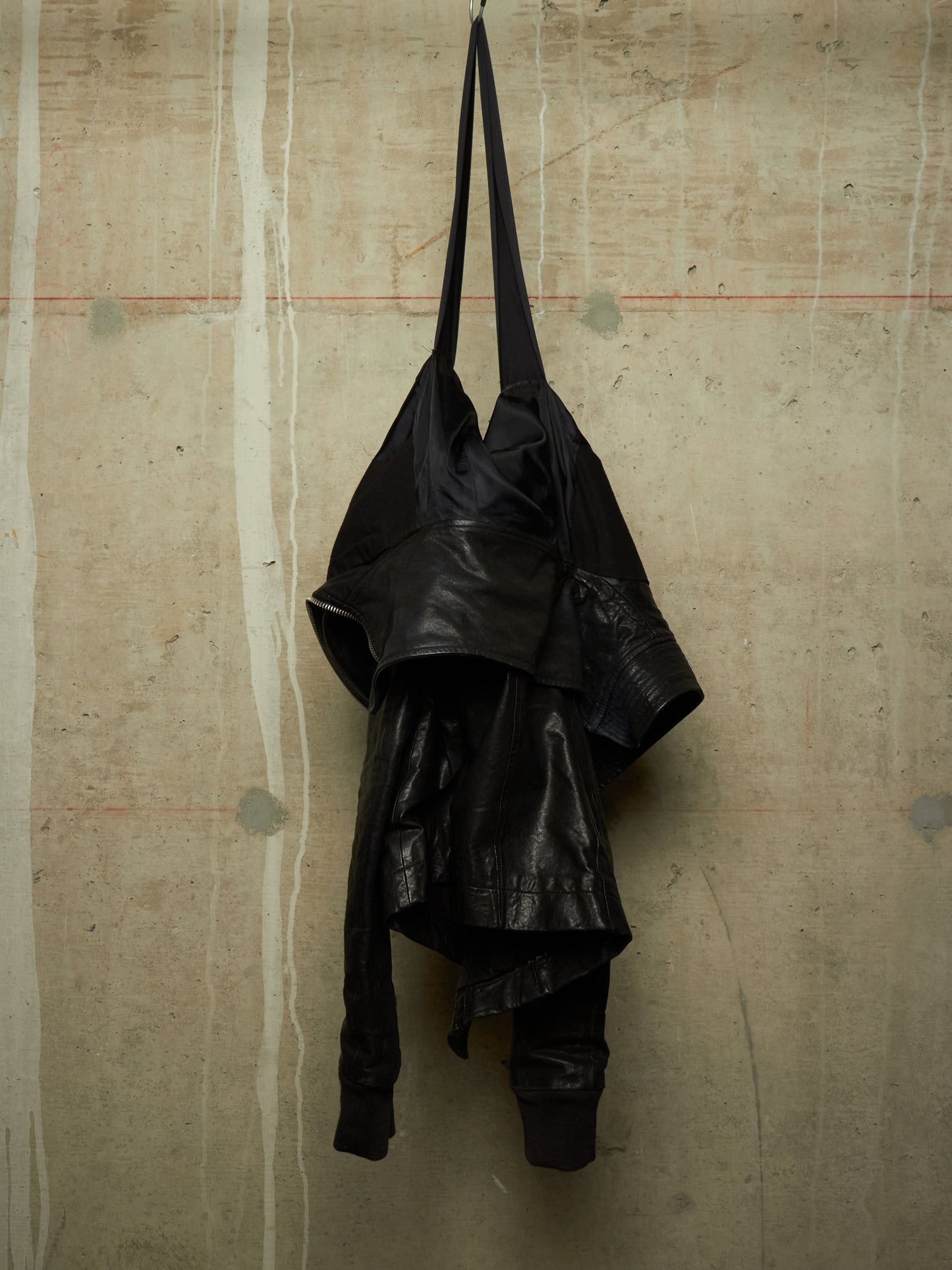 A black leather shoulder bag with a long strap, hanging against a textured concrete wall.