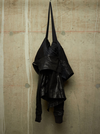 A black leather shoulder bag with a long strap, hanging against a textured concrete wall.