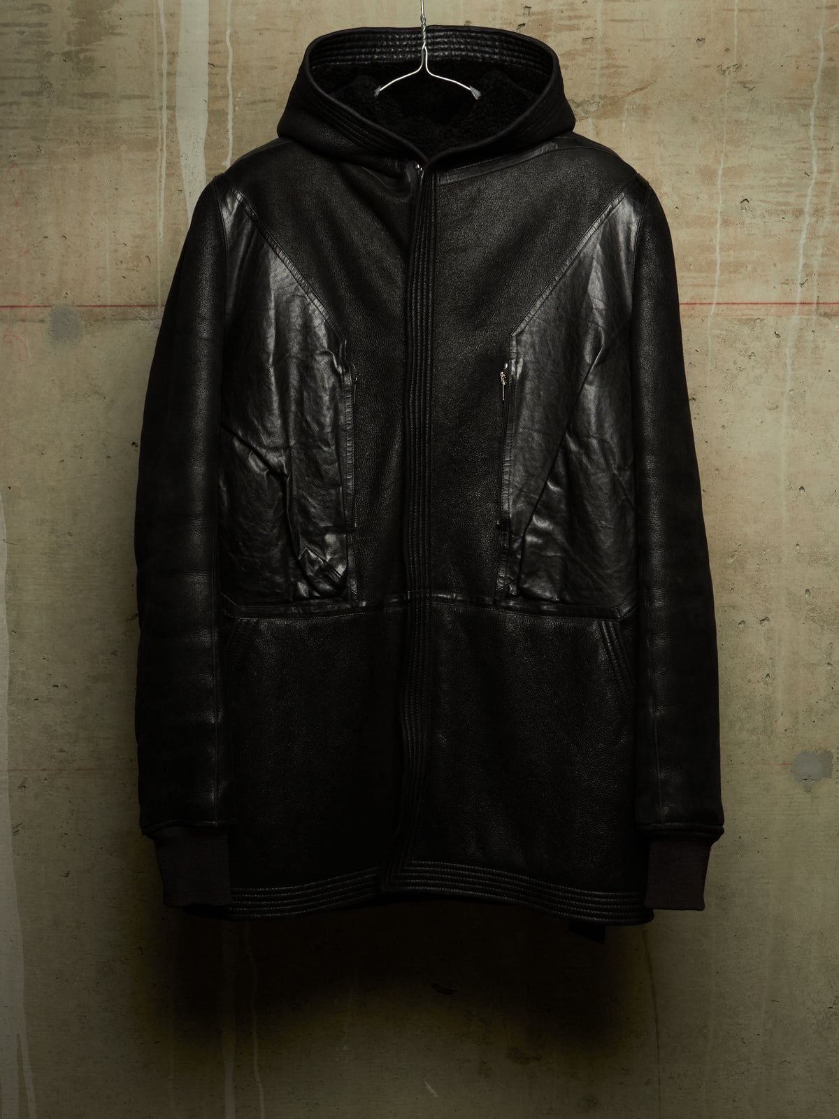A black leather hooded jacket with a front zipper and zippered pockets, hanging on a wire hanger against a textured concrete wall.