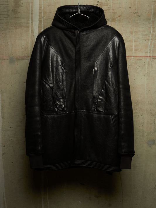 A black leather hooded jacket with a front zipper and zippered pockets, hanging on a wire hanger against a textured concrete wall.