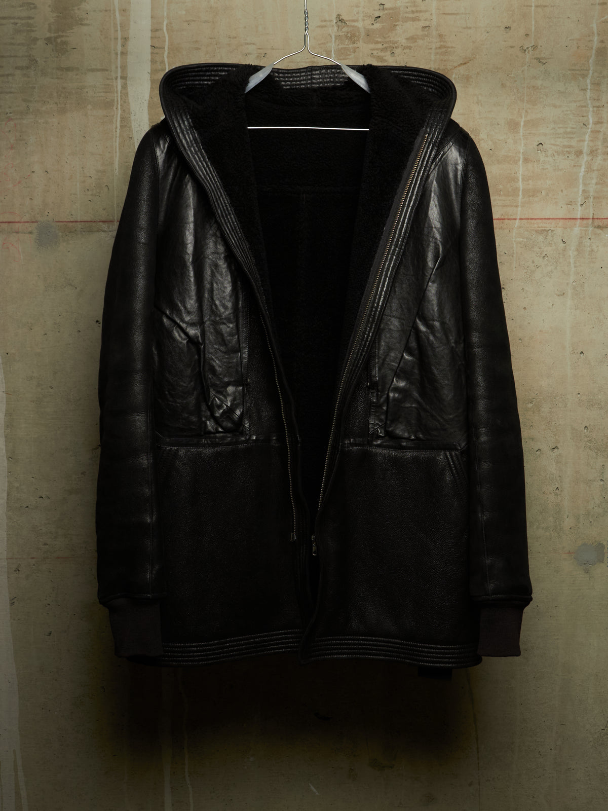 A black leather hooded jacket with ribbed trim and a black fleece-lined interior, hanging on a wire hanger against a textured concrete wall.