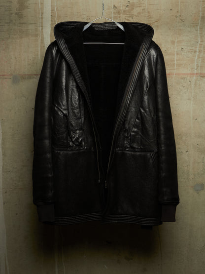 A black leather hooded jacket with ribbed trim and a black fleece-lined interior, hanging on a wire hanger against a textured concrete wall.