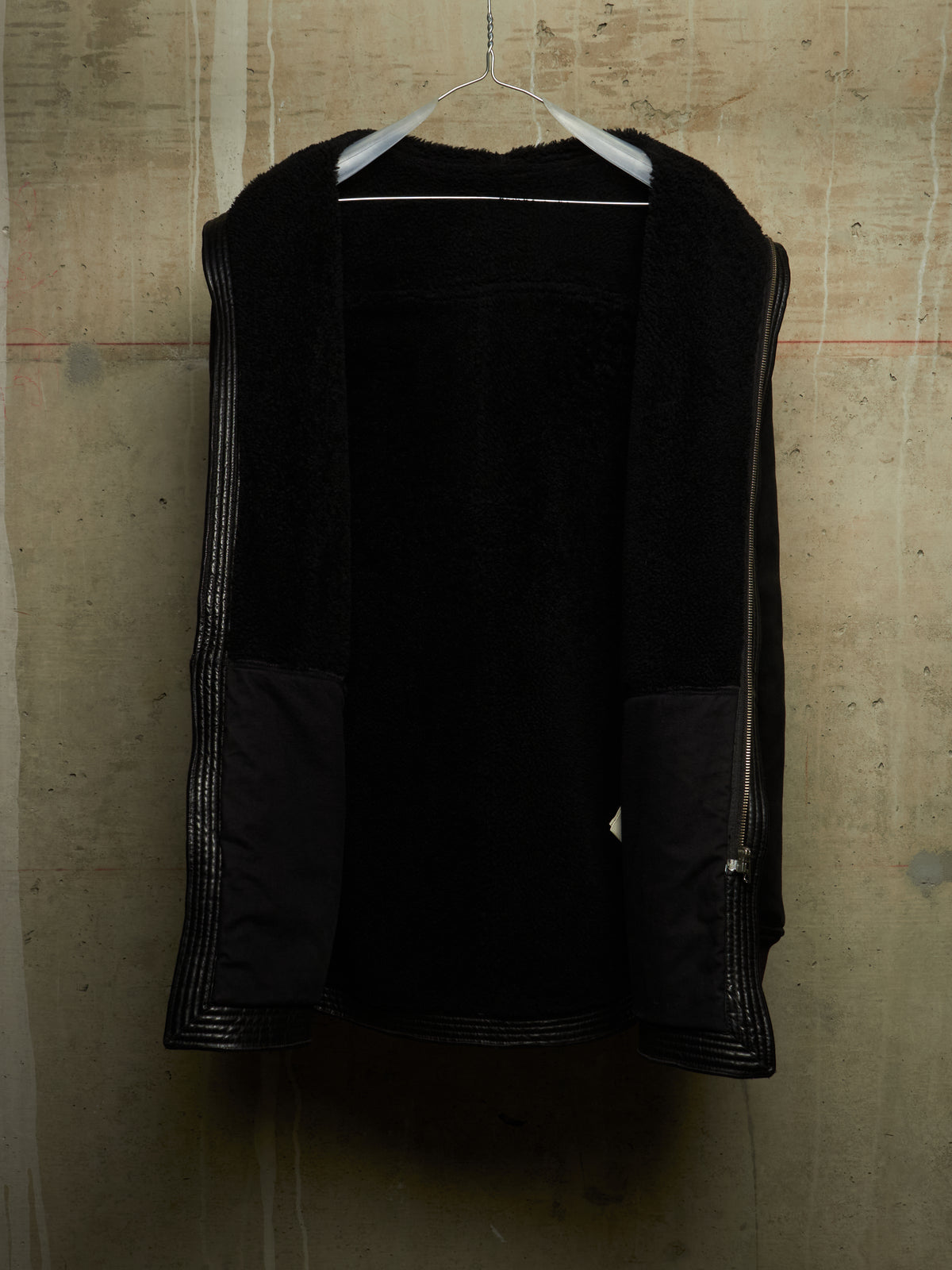 A black open-front jacket with textured fuzzy lining and silver-toned zipper details, hanging on a white hanger against a concrete wall.
