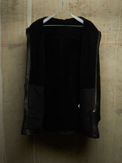 A black open-front jacket with textured fuzzy lining and silver-toned zipper details, hanging on a white hanger against a concrete wall.