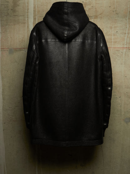 A black leather hooded jacket shown from the back, hanging against a textured concrete wall.