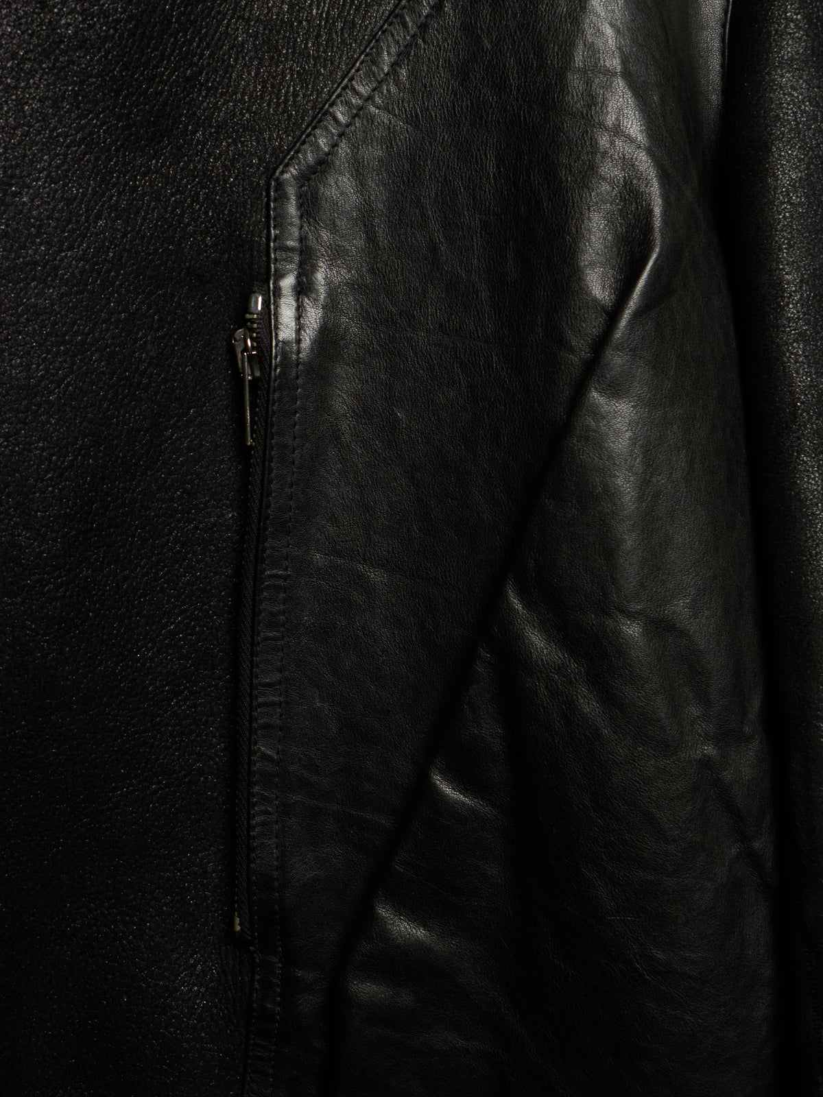 Close-up of a black leather jacket showing the textured leather material, a diagonal seam, and a dark metal zipper.