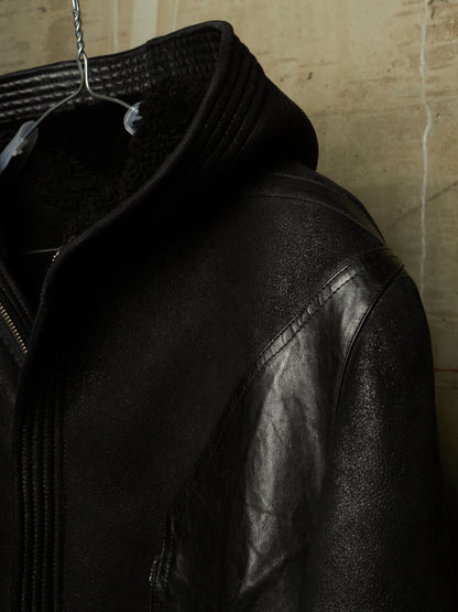 A close-up of a black leather jacket with a high ribbed collar and a plush black fleece lining, hanging on a metal hanger against a textured beige wall.