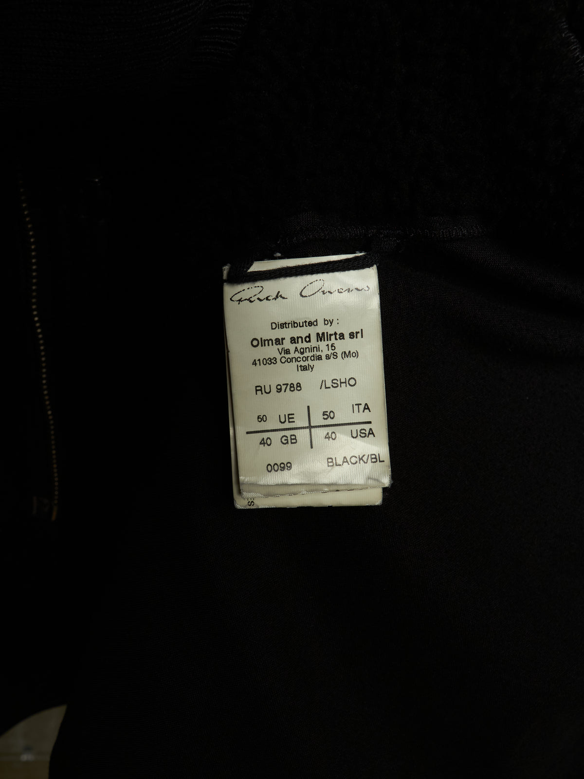 A close-up of a black garment showing a sewn-on size and brand label that reads 'Greek Oven' and 'Distributed by Olmar and Mirta art'.