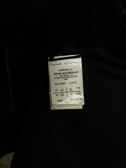 A close-up of a black garment showing a sewn-on size and brand label that reads 'Greek Oven' and 'Distributed by Olmar and Mirta art'.
