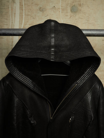 A black leather hooded jacket with a high ribbed hood and front zipper, hanging against a textured beige wall.