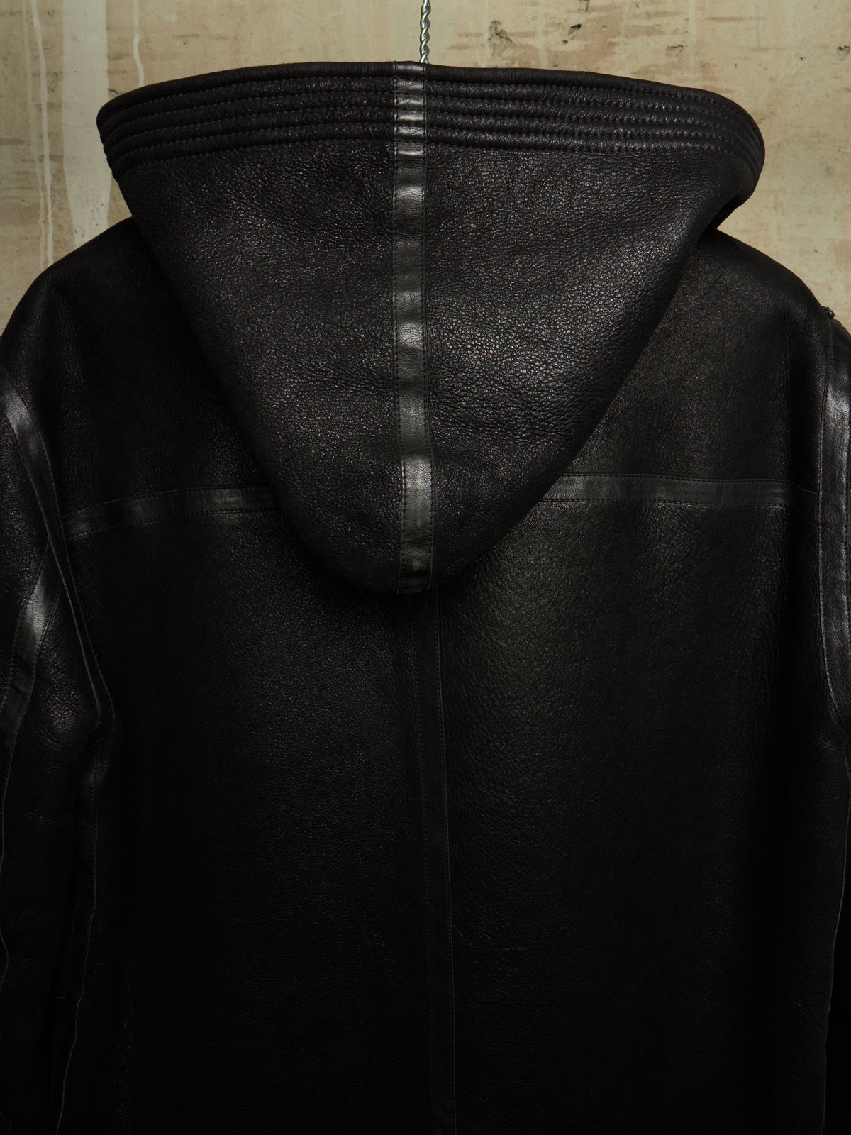 Close-up of the back of a black leather hooded jacket, showing the hood, stitched seams, and textured leather material against a neutral background.