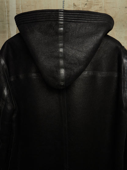 Close-up of the back of a black leather hooded jacket, showing the hood, stitched seams, and textured leather material against a neutral background.