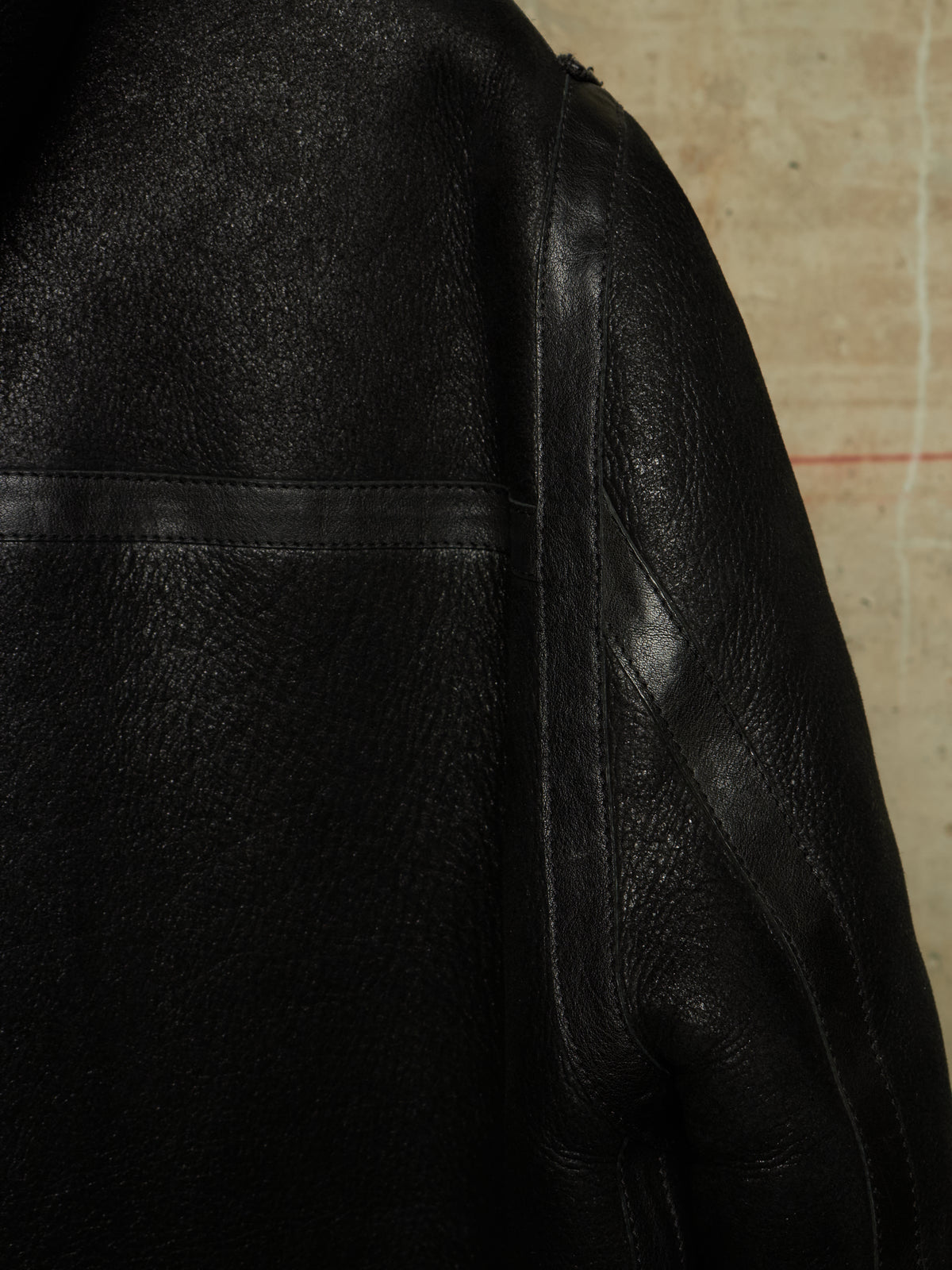 Close-up of a black leather jacket showing the textured grain, stitching, and shoulder seam against a neutral beige background.