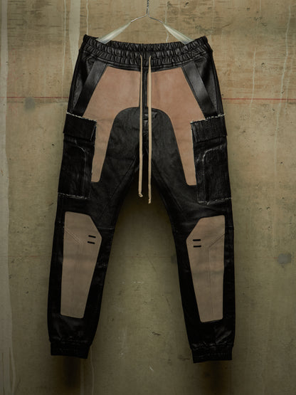 A pair of black and beige patchwork cargo jogger pants hanging against a textured concrete wall. The pants feature a mix of matte and glossy black panels with large beige patches, a drawstring waist, and side cargo pockets.