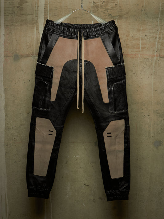 A pair of black and beige patchwork cargo jogger pants hanging against a textured concrete wall. The pants feature a mix of matte and glossy black panels with large beige patches, a drawstring waist, and side cargo pockets.