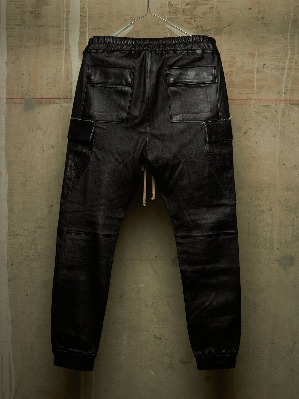 Black leather cargo pants with an elastic waistband and drawstring, shown from the back hanging against a textured concrete wall.