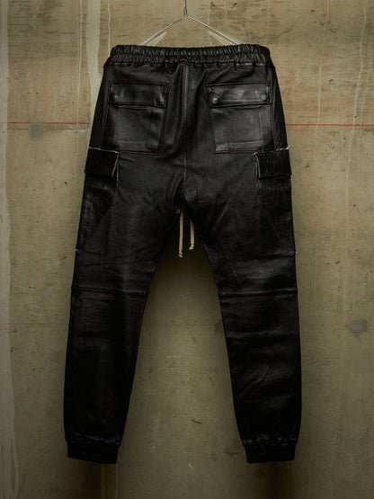 Black leather cargo pants with an elastic waistband and drawstring, shown from the back hanging against a textured concrete wall.