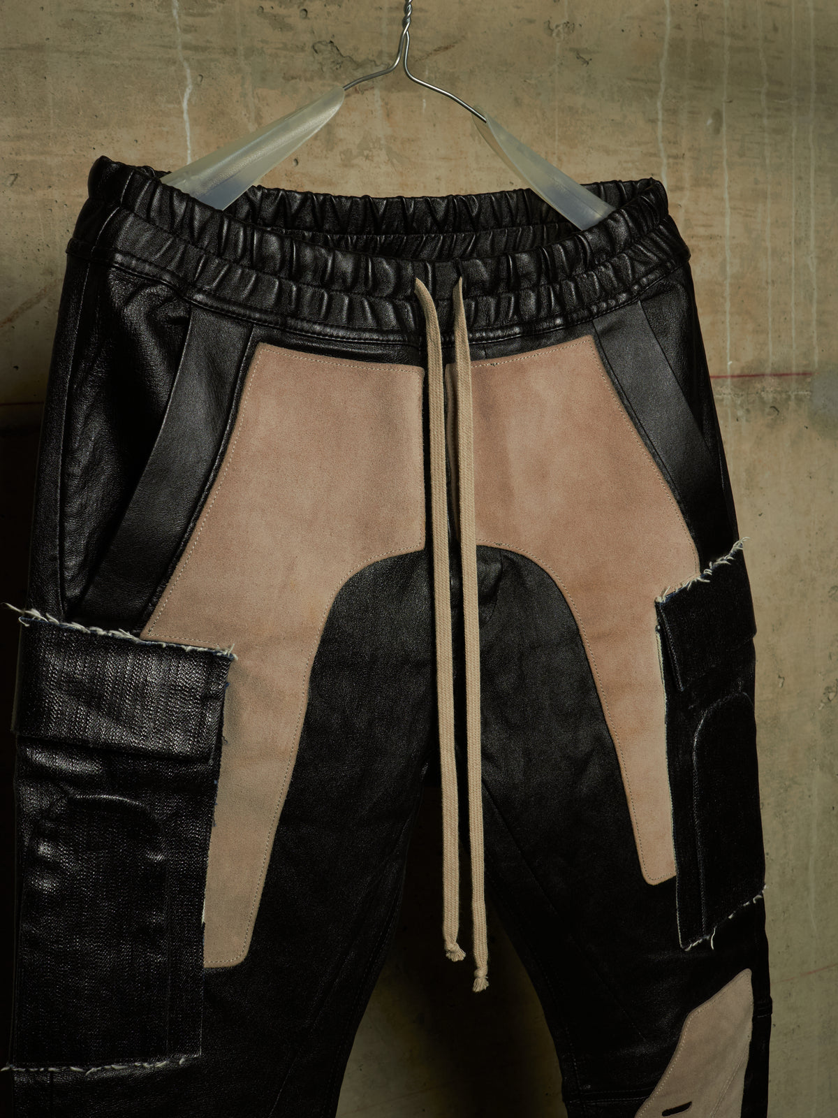 Patchwork cargo pants made of black leather and beige suede, hanging on a metal hanger against a textured concrete wall.