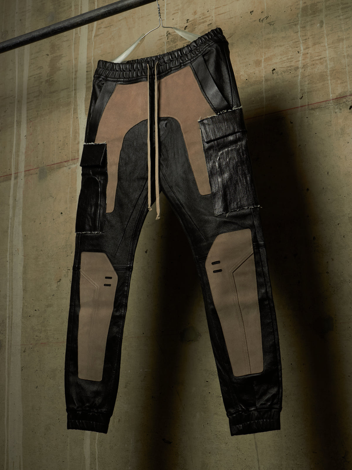 Black leather cargo jogger pants with tan suede-like panel inserts, hanging on a white hanger against a textured concrete wall.