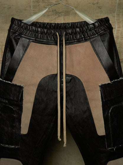 Black and beige patchwork cargo pants with a drawstring waist, hanging on a metal hanger against a textured concrete wall.