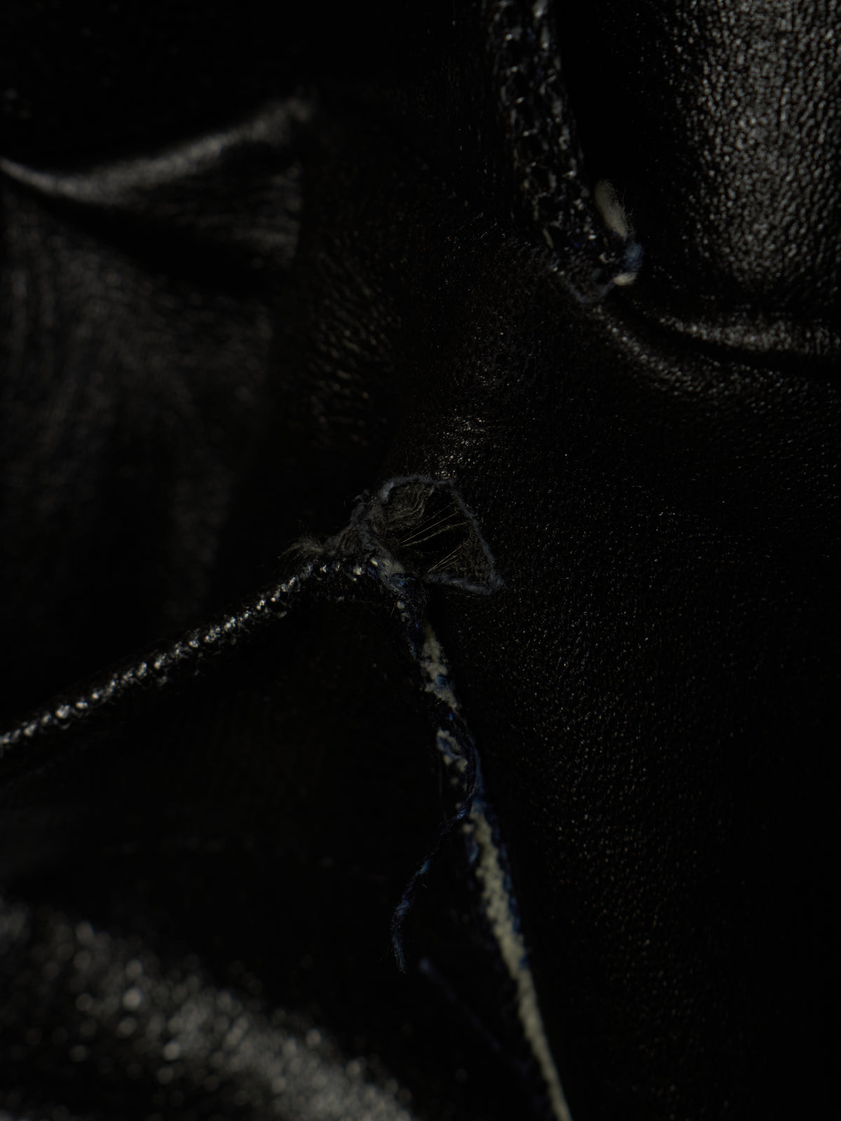 Extreme close-up of a black leather boot or shoe showing the textured grain, a stitched seam, and frayed thread details in very dark lighting.