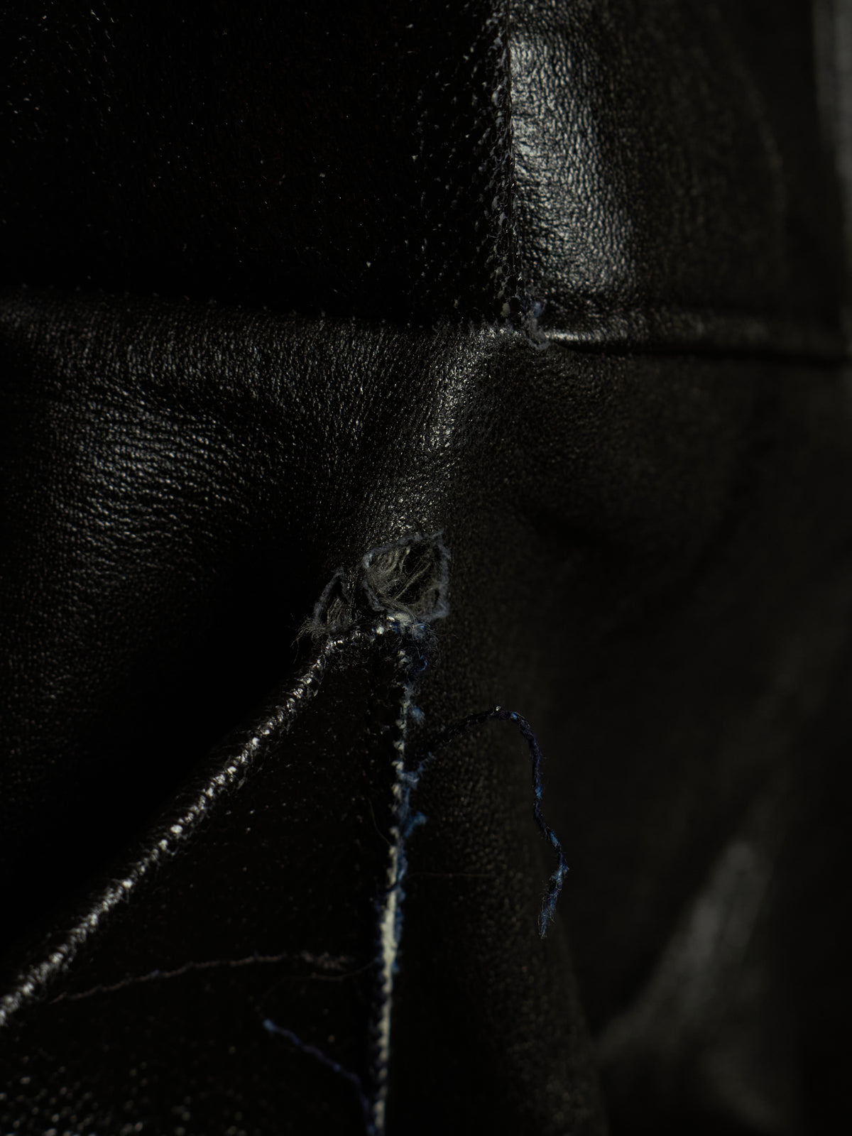 Close-up of a black leather item showing a small tear or hole near the center, with visible thread and dark textured material around it.