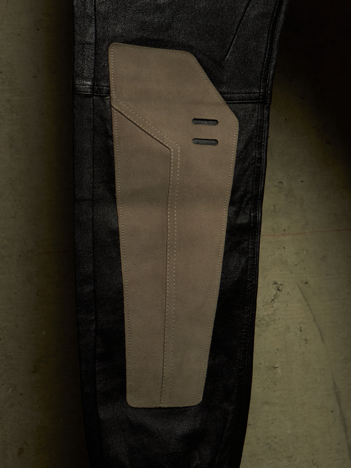 Close-up of a pair of dark washed jeans with a large tan suede or nubuck patch panel on the upper thigh, set against a dark concrete floor.