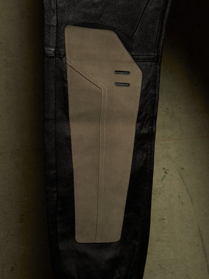 Close-up of a pair of dark washed jeans with a large tan suede or nubuck patch panel on the upper thigh, set against a dark concrete floor.