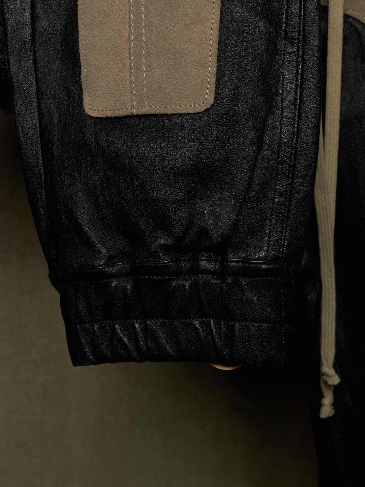 Close-up detail of a dark washed denim jacket showing a tan suede patch pocket and a ribbed elastic cuff.