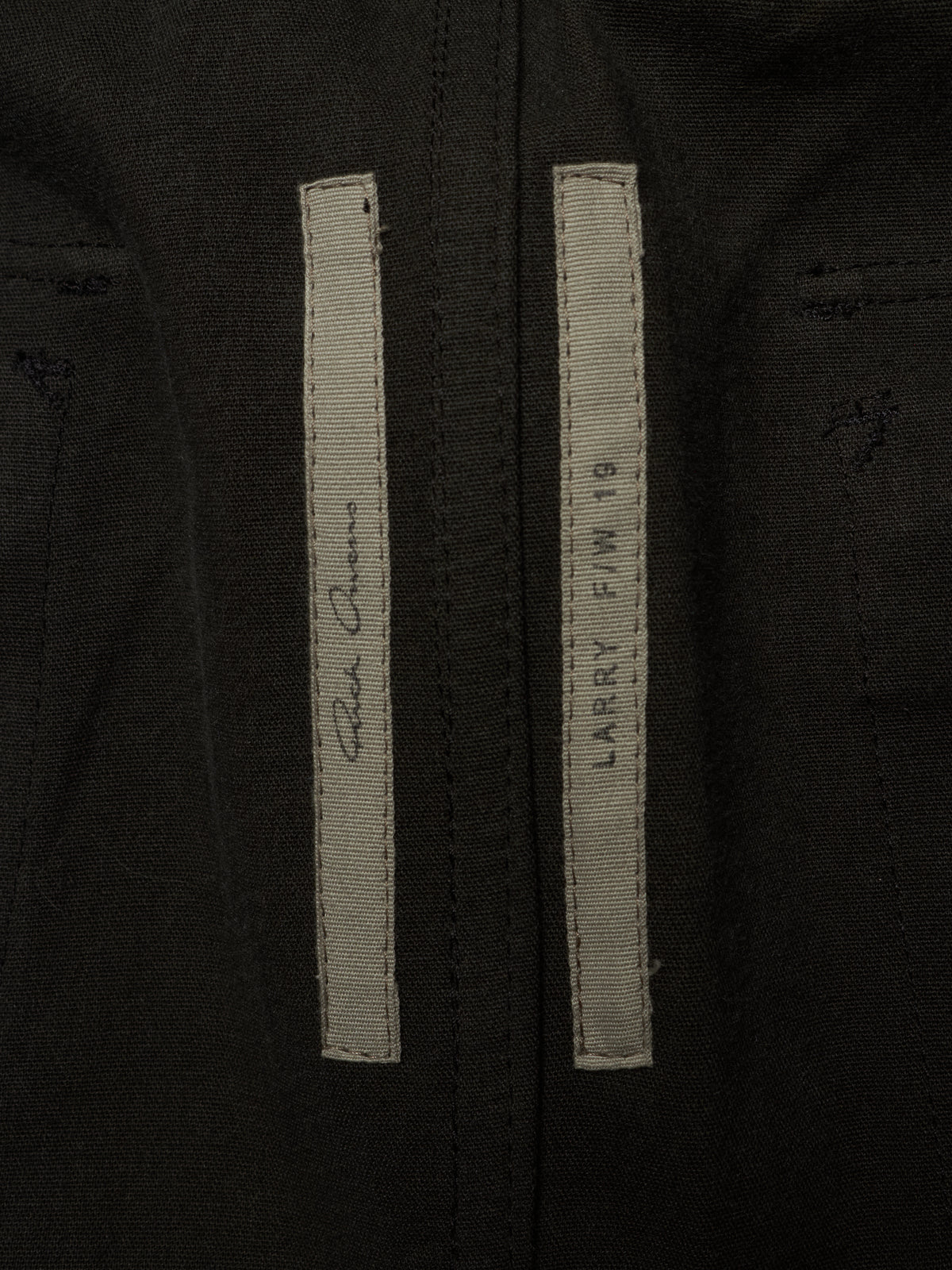 Close-up of a black jacket or coat showing two vertical light gray labels stitched on the front, with one label featuring a signature and the other reading 'LARRY F/W 18' and 'SANTA MONICA'.