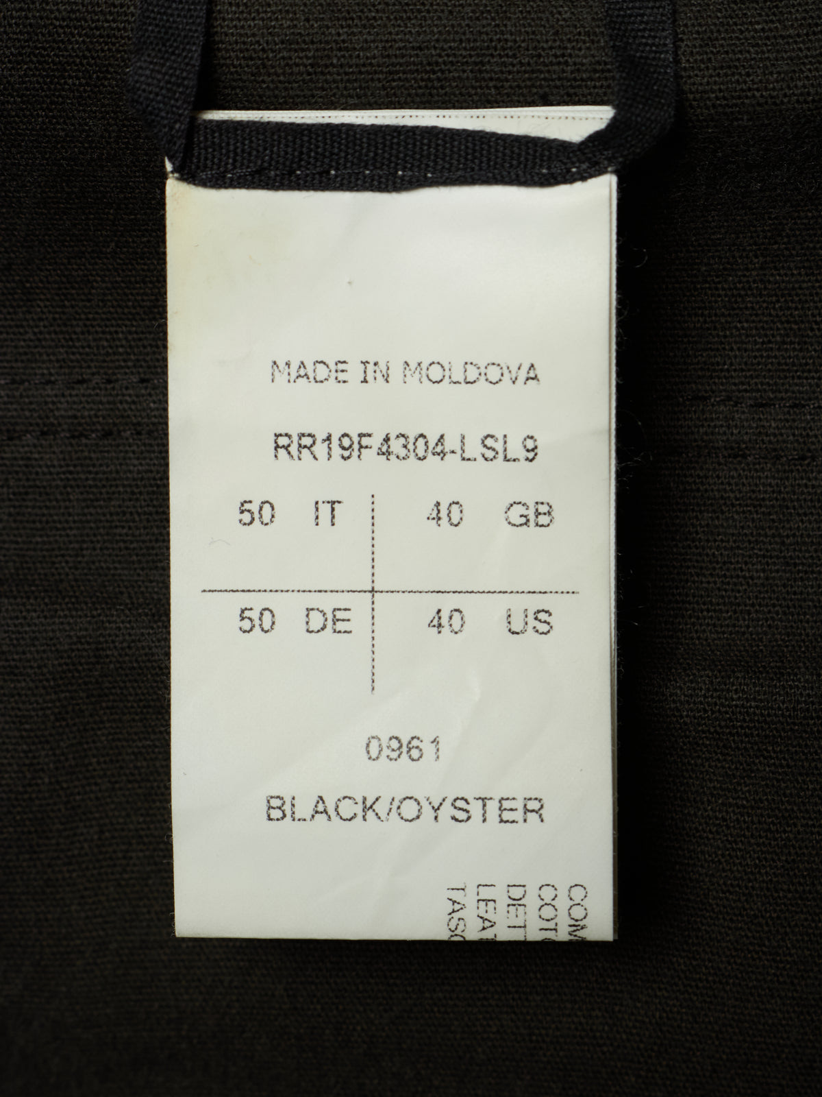 A close-up of a white paper garment tag sewn onto a black fabric item. The tag shows printed text including 'MADE IN MOLDOVA', a product code, weight and size markings, and 'BLACK/OYSTER' at the bottom.